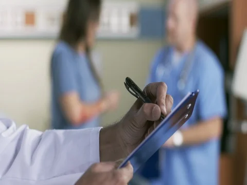 Hand writes notes in foreground as male nurse treats patient in emergency room 4 Stock Footage 69541448