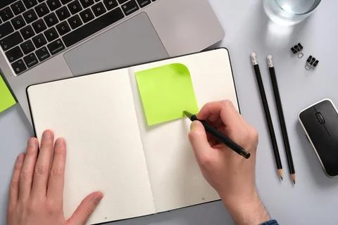 Hand writing on a green colored post-it note above an open notepad on a desk. Stock Photos