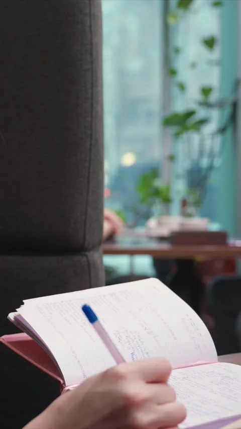 Hand Writing in Notebook Close Up with Soft Natural Light and Blurred Plant Stock Footage 348162560