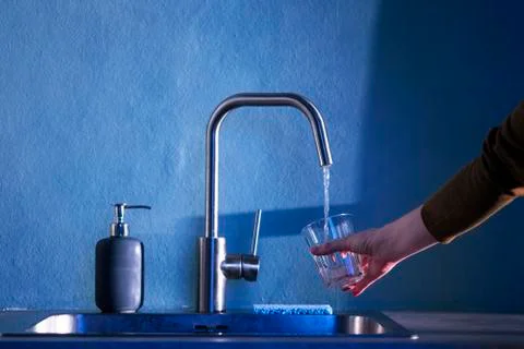 The hand of a young woman pours clean water from under a kitchen faucet into Stock Photos