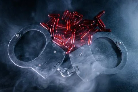 Handcuffs resting on a dark surface, surrounded by red capsules, with smoke.. Foto stock