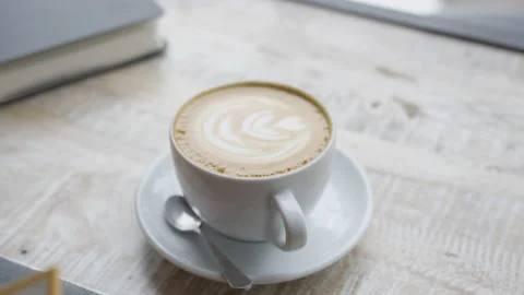 Handheld close up of cup of coffee with latte art foam Sitting On wooden Table Stock Footage 208724199