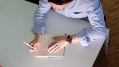 Handheld overhead view of young man sitting at desk and writing in notebook Vídeo Stock 120426189