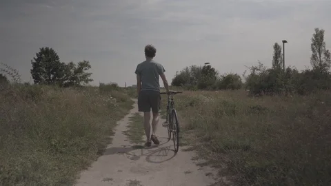 Handheld shot of a boy walking along with his cycle on the dirt path Stock Footage 114881154