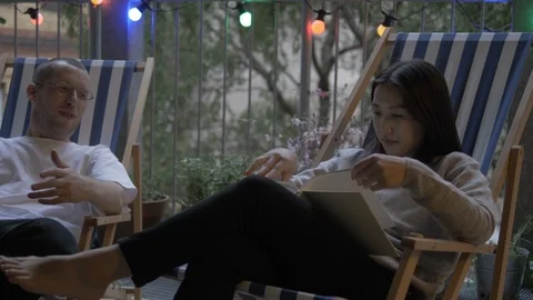Handheld shot of couple drinking wine and reading book while sitting on chair Stock Footage 114842945