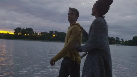 Handheld shot of happy couple talking while walking at riverbank during sunset Stock Footage