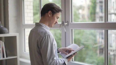 Handheld shot of man reading book standing in front of window Stock Footage 114877953