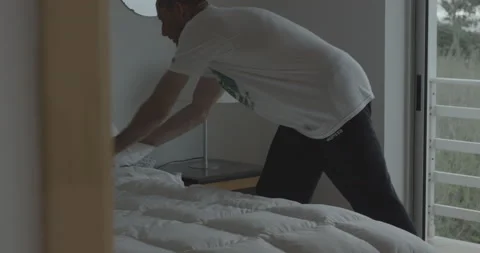 Handheld shot of pro surfer David Langer making his hotel bed neater in morning Stock Footage 160110199
