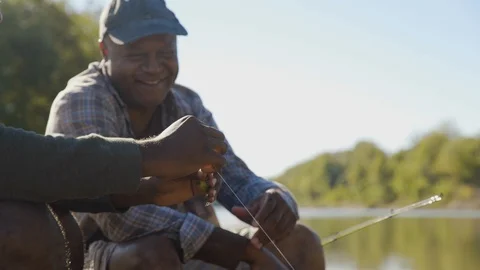 Handheld shot of smiling father guiding son in tying fishing bait to rod while Stock Footage