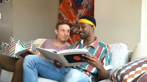 Handheld shot of smiling friends looking picture books while sitting on sofa 스톡 동영상 114866482