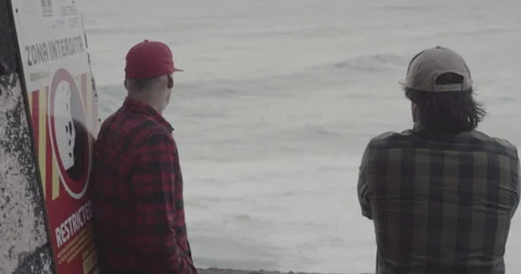 Handheld shot of two pro surfers standing on edge off cliff looking at ocean Stock Footage 160114201