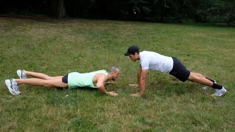 Handheld shot of two young men in sportswear doing push-ups together on grass Stock Footage 114879634