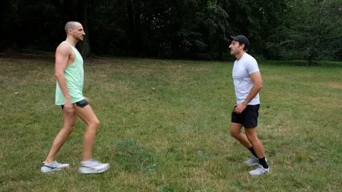 Handheld shot of two young men talking while doing lunges together on grass Stock Footage 114879646