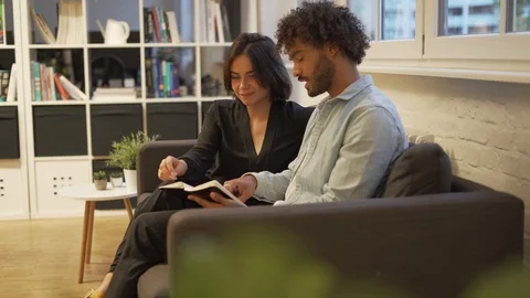 Handheld shot of young couple talking and reading book while sitting on sofa Stock Footage 114874926