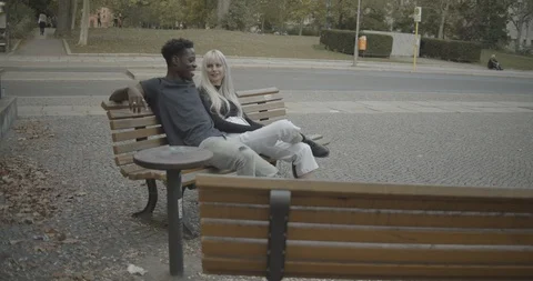 Handheld shot of young couple talking while sitting on park bench by road Stock Footage 120429312