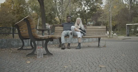 Handheld shot of young couple talking while sitting on park bench by road Stock Footage 120430534