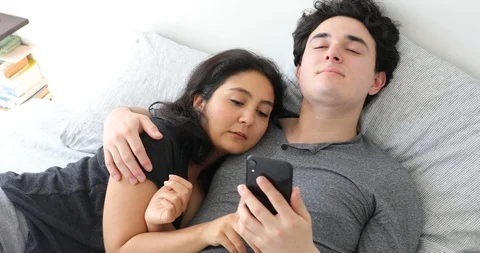 Handheld shot of young couple using mobile phone while lying down in bed Stock Footage 120457628
