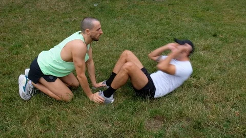 Handheld shot of young man helping another doing sit-ups on lawn Stock Footage 114879588