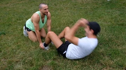 Handheld shot of young man helping another doing sit-ups on lawn Stock Footage 114879593