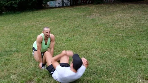 Handheld shot of young man helping another doing crunches on lawn Vídeo Stock 120426302