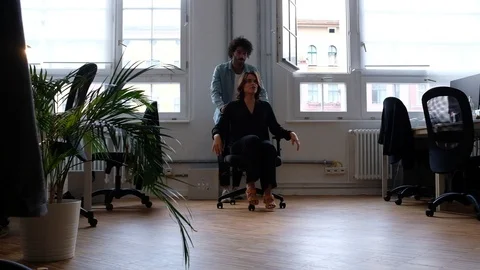 Handheld shot of young man pushing young woman on chair through office space 스톡 동영상 120419030