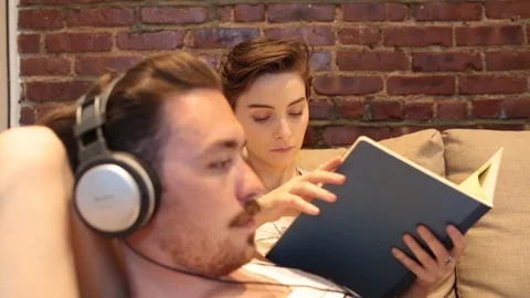 Handheld shot of young man using laptop while woman reading book on sofa Video stock 114862085