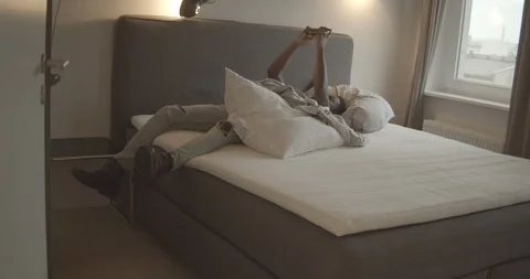 Handheld shot of young man using smartphone and lying on bed in bedroom Stock Footage 120423053