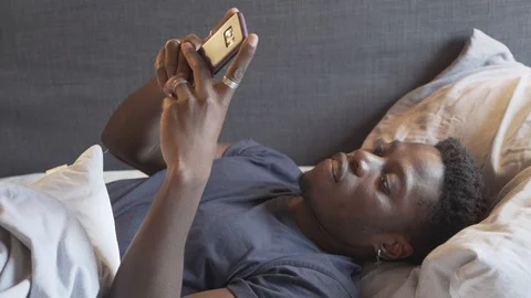 Handheld shot of young man using smartphone while lying on bed in bedroom Stock Footage 120426116