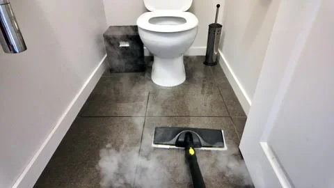 A handheld steam mop cleans grey tiled bathroom flooring around a white toile Stock Footage 328889527