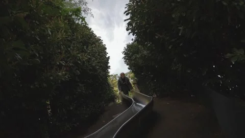 Handheld View of Blonde Man Sliding Down Playground Slide in London Park Vídeo Stock 336738174