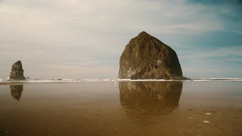 Handheld wide low angle of Haystack Rock with waves and seagulls walking Stock Footage 102418203