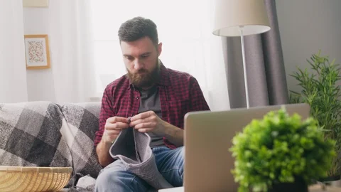 Handheld of young man sitting on sofa and watching knitting tutorial on laptop Video stock 107352741