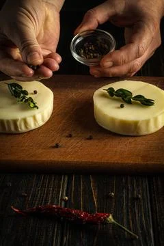 Hands add spices to two cheese rounds placed on a wooden cutting board. The b Stock Photos