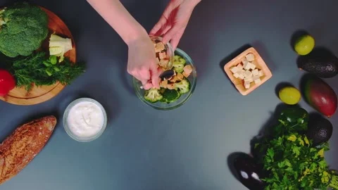 Hands adding cubed tofu to broccoli salad on kitchen table overhead Vidéo 342866858