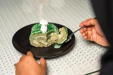 Hands and a fork cut into a slice of green pandan cake roll on a black plate. Foto stock