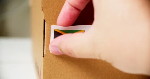 Hands applying MADE IN JAMAICA flag label on a shipping cardboard box with Stock Footage 201019340