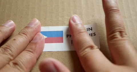Hands applying MADE IN PHILIPPINES flag label on a shipping box with Stock Footage 253381979