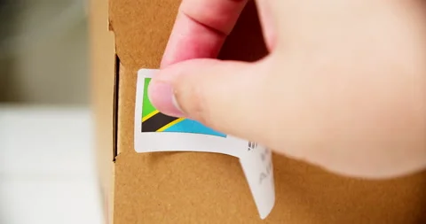 Hands applying MADE IN TANZANIA flag label on a shipping cardboard box Stock Footage 201111972