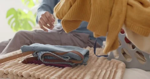 Hands are folding and stacking clothes on bed by window, starting with burgundy Stock Footage 347309504