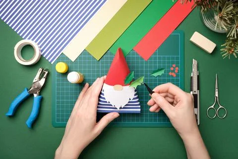 Hands assembling a DIY Christmas gnome from colored paper on a green cutting  Stock Photos