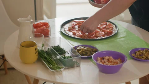 Hands assembling tomato pizza on round metal tray with bowls of cheese, olives Stock Footage 343682414