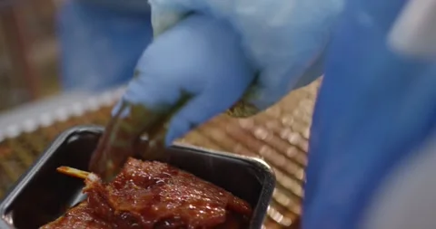 Hands in blue gloves placing glazed marinated meat into a dark tray in a food Vídeo Stock 339999291
