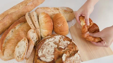 Hands Breaking Fresh Croissant Beside Assorted Bakery Breads on Wooden Board Stock Footage 337746760