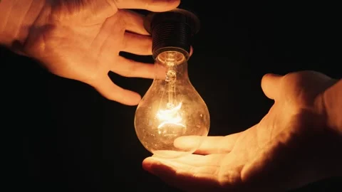 Hands Carefully Holding Glowing Lightbulb with Warm Light in Darkness Stockbeeldmateriaal 334128712