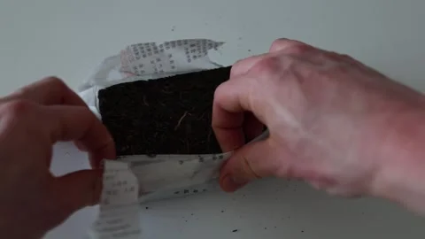Hands carefully unwrapping a compressed tea brick from its traditional paper pac Stock Footage 304623455