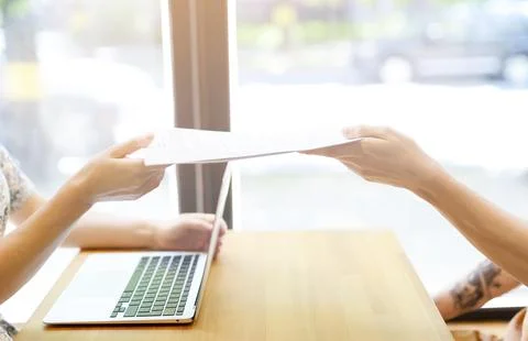 Hands in close-up pass documents to each other above the table with the laptop. Stock Photos