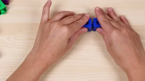 Hands Connecting Blue and Yellow Interlocking Plastic Cubes on Table Stock Footage 326019596