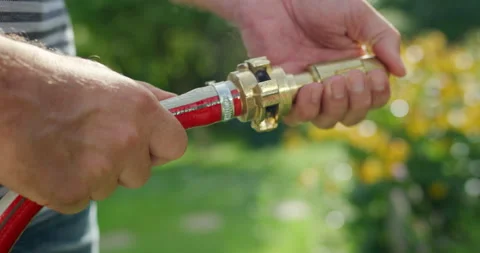 Hands connecting a brass coupling to a red garden hose in a sunny garden Video stock 329289098