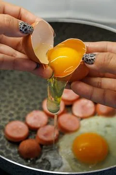 Hands crack egg into frying pan with cooked sausages Stock Photos