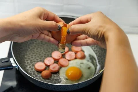 Hands crack egg over sauteing sausage slices Stock Photos